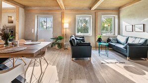 Een ruime woonkamer met een houten eettafel, een bank en een zithoek. Grote ramen zorgen voor natuurlijk licht en uitzicht op de omgeving.