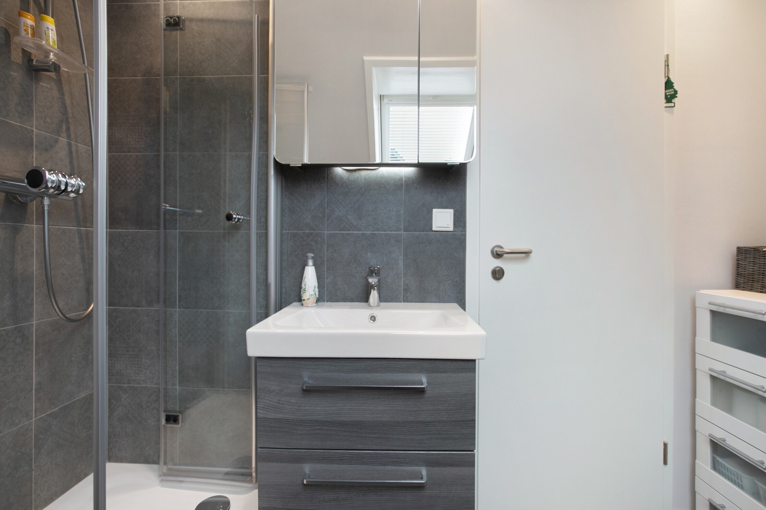 A modern bathroom with a glass shower enclosure, a white sink with a mirror above, and a built-in cabinet with drawers.