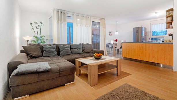 Ein modernes Wohnzimmer mit einem braunen Ecksofa, einem Holztisch und einer Küchenzeile mit Holzschränken und einem Edelstahlkühlschrank.
