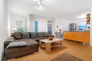 Un salon moderne avec un canapé d’angle marron, une table basse en bois et un coin cuisine doté de meubles en bois et d’un réfrigérateur en acier inoxydable.