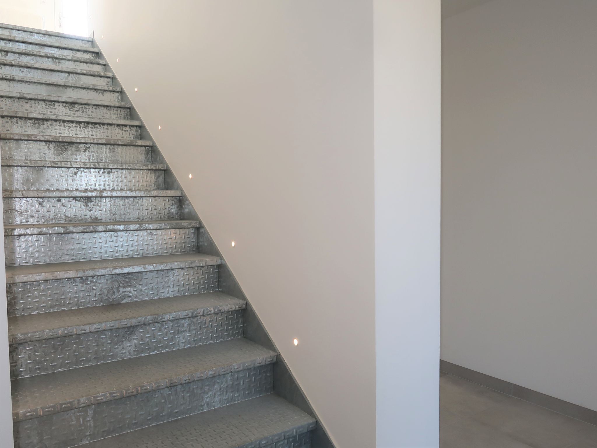 Una escalera con acabado metálico texturizado y una pared blanca lisa a su lado.