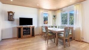 A dining area with a wooden table and chairs, a television on a cabinet, and a window with curtains. - Triberg-Blick Apartment with Mountain View, Terrace and Wi-Fi (Triberg im Schwarzwald)