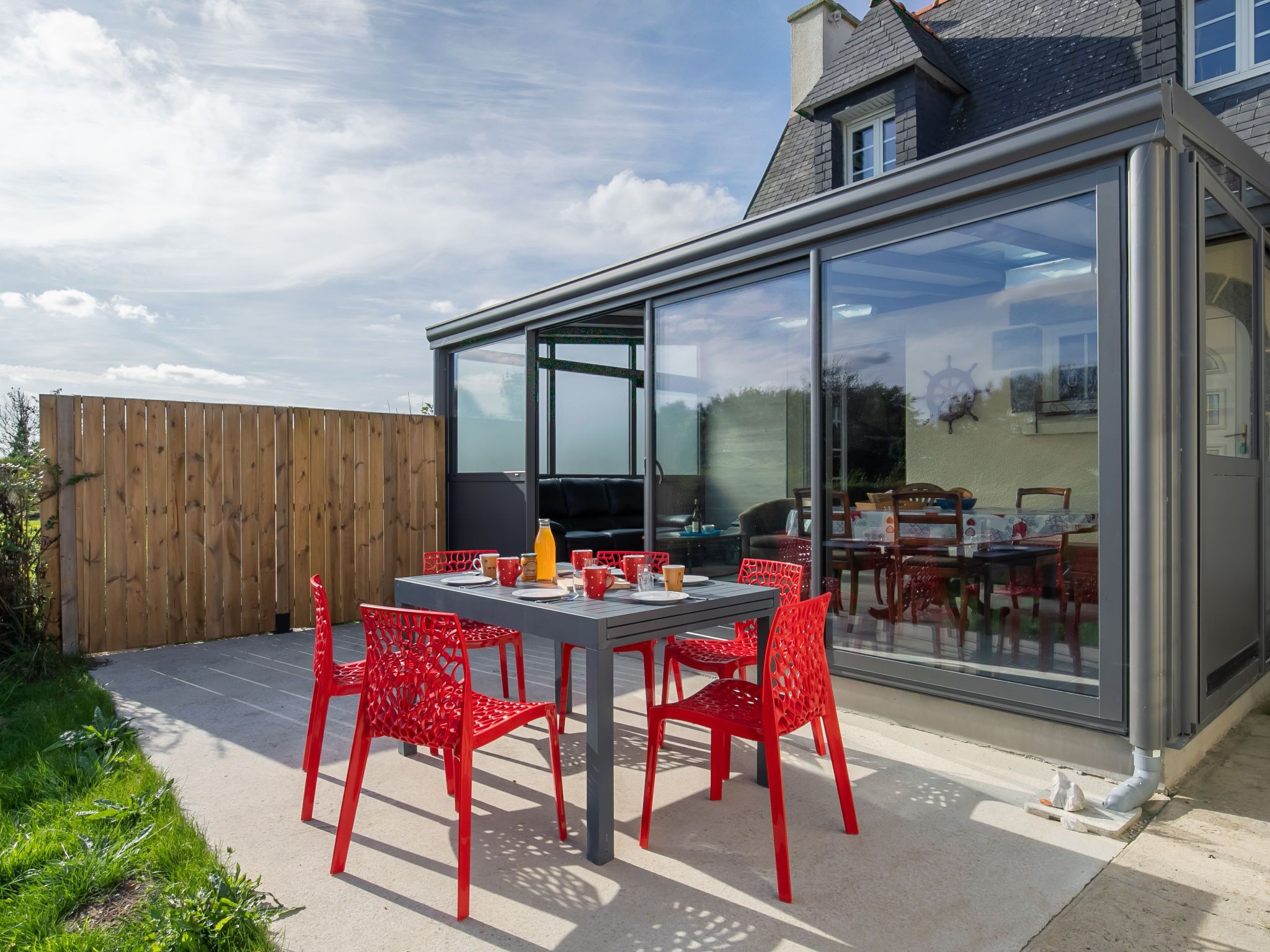Une terrasse extérieure avec une table dressée pour quatre personnes, entourée de chaises rouges, et une véranda fermée par des baies vitrées donnant sur le jardin.