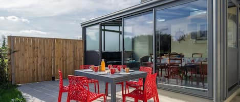 Een terras buiten met een tafel voor vier personen, omringd door rode stoelen, en een glazen overkapping met uitzicht op de achtertuin.