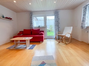 A room with a red sofa, a wooden floor, a white chair, and a window with curtains.