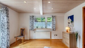 Ein Zimmer mit Holzboden, Heizkörper, Sessel, Schreibtisch mit Schubladen, Vorhängen am Fenster und einem Bild an der Wand.