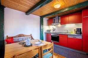A wooden dining table with chairs, a red kitchen with appliances, and a wooden ceiling.