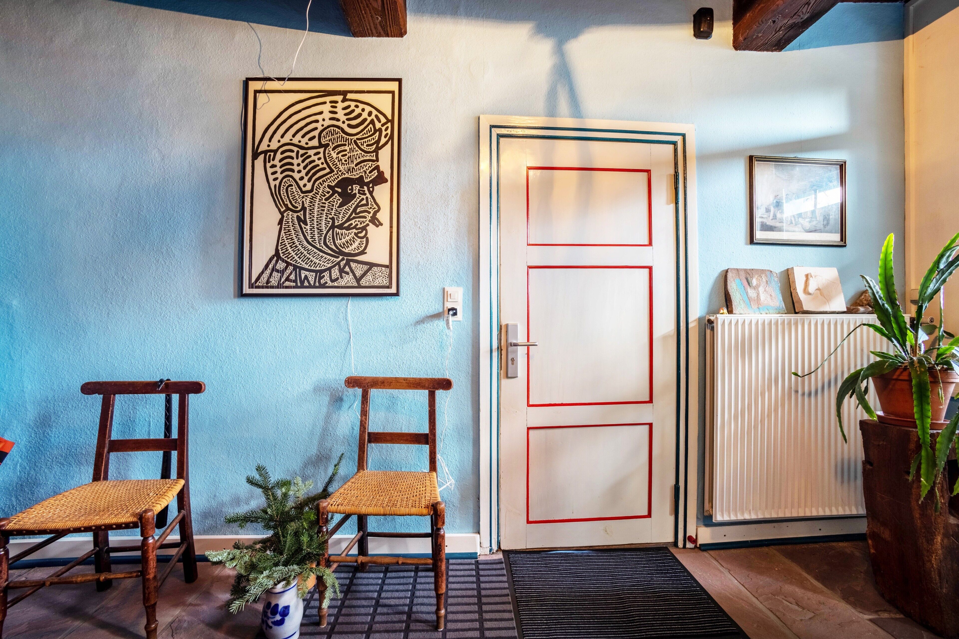 Une pièce avec deux chaises en bois, une porte dont le cadre est rouge, un radiateur et un tableau encadré accroché au mur.