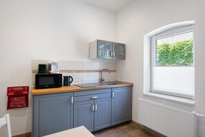Une cuisine compacte équipée d’un four à micro-ondes, d’une machine à café et d’un évier. Il y a un placard au-dessus de l’évier et une fenêtre avec des stores.