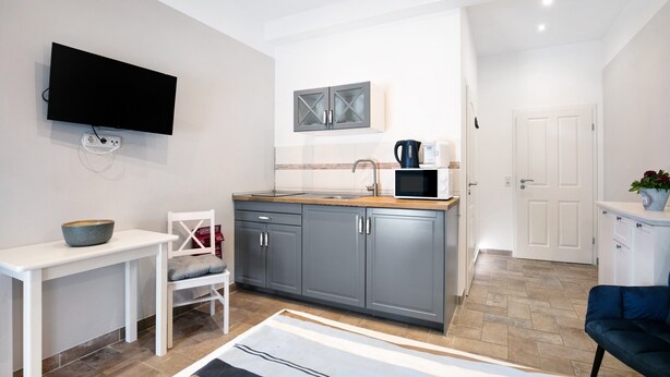 A modern kitchen with grey cabinets, a white countertop, a microwave, and a kettle. There is a white table with a bowl on it and a chair with a cushion. A flat-screen TV is mounted on the wall. - Studio Apartment 'Appartement 2 - Galopp' with Shared Terrace and Wi-Fi (Ludwigsfelde)