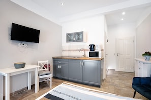A modern kitchen with grey cabinets, a white countertop, a microwave, and a kettle. There is a white table with a bowl on it and a chair with a cushion. A flat-screen TV is mounted on the wall.