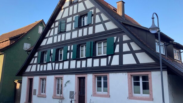 Una casa tradicional de entramado de madera con techo a dos aguas pronunciado, múltiples ventanas con contraventanas y un pequeño porche con escalones.
