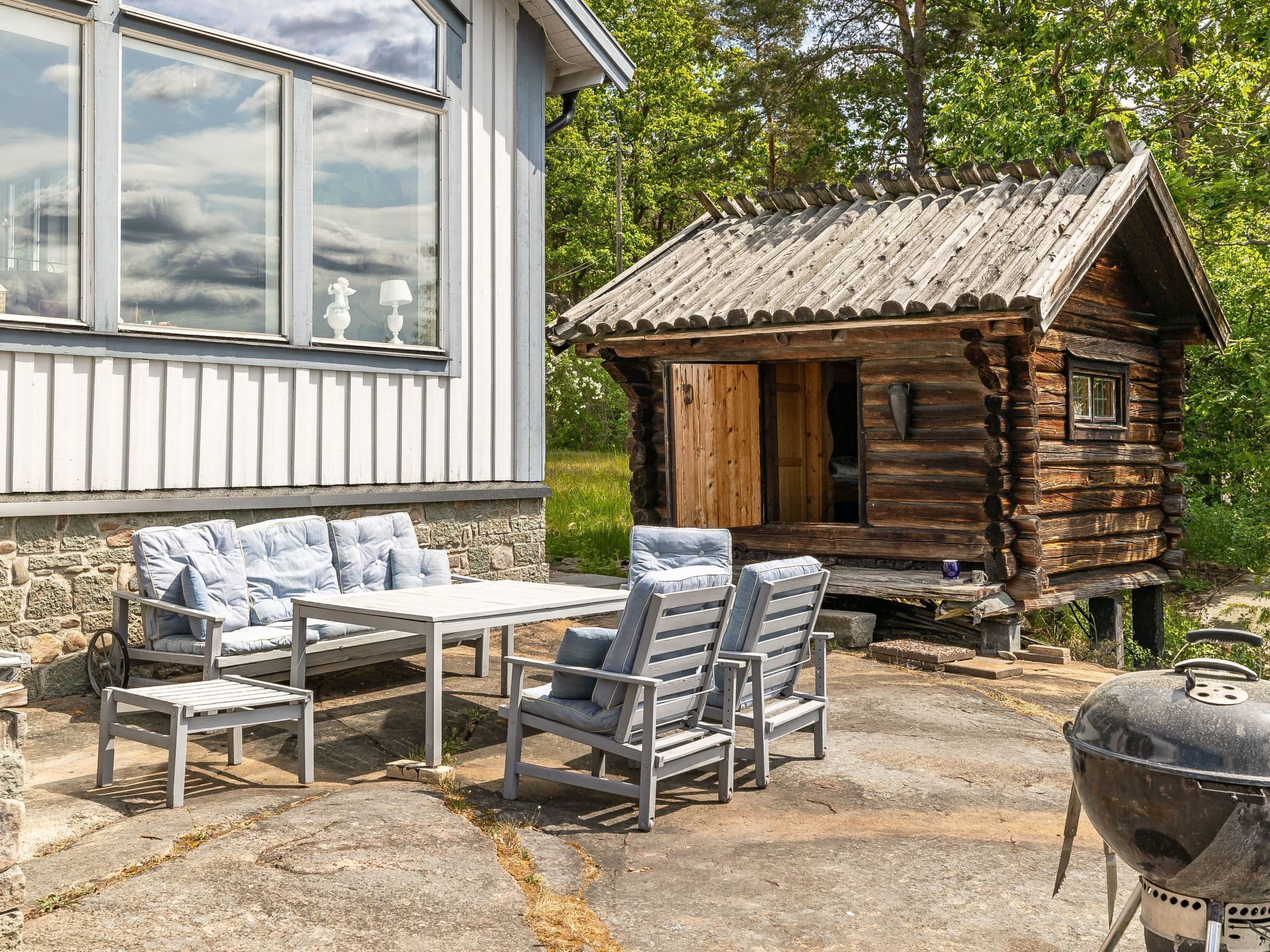 A log cabin with a stone patio, a barbecue grill, and a white building with large windows.
