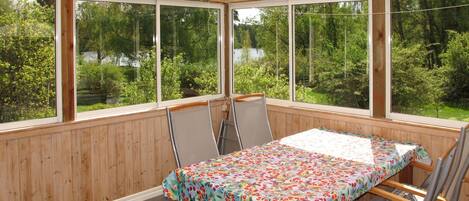 A sunlit patio with a table and chairs, large windows offering a view of a lake and forest.