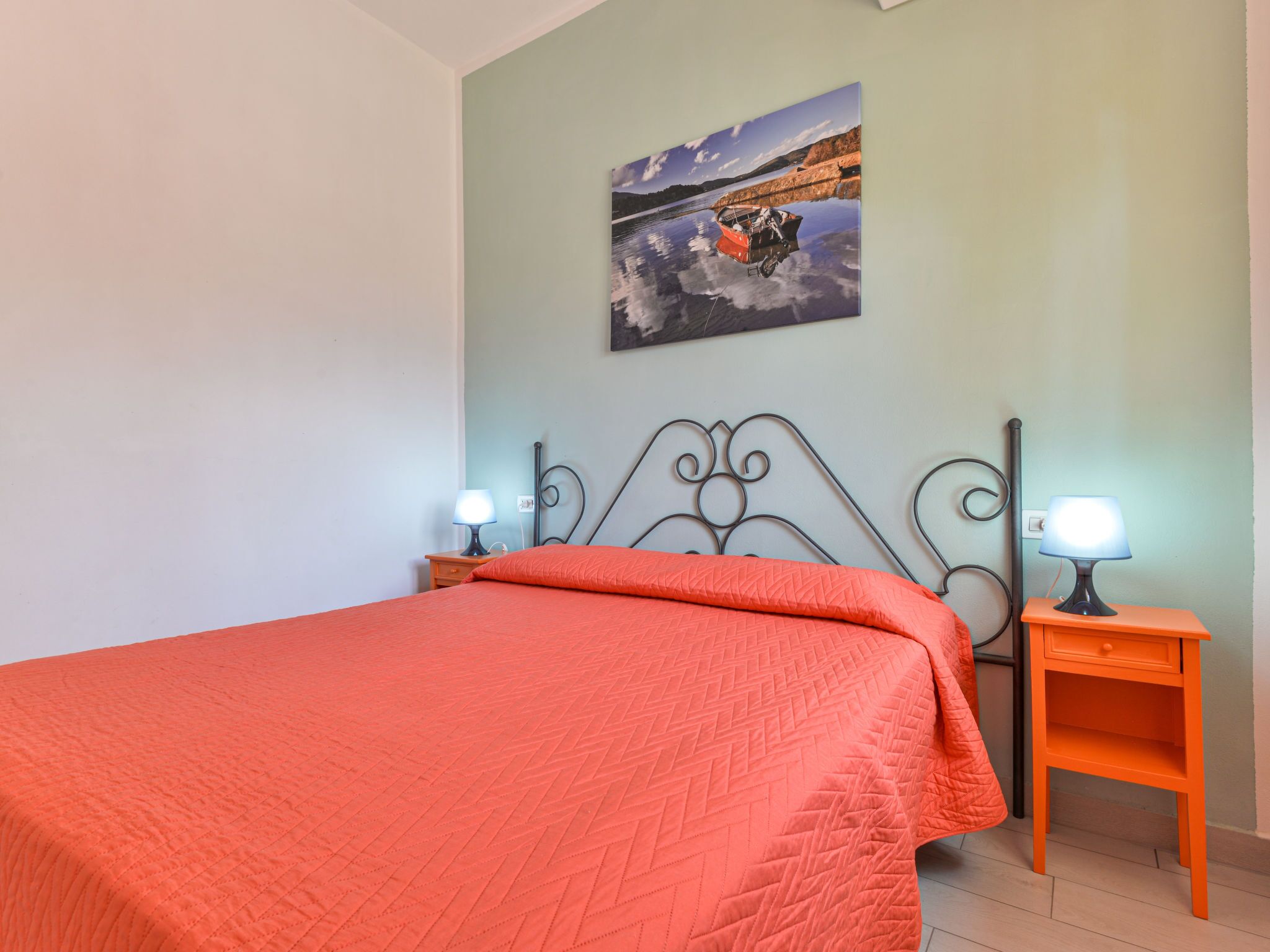 Quarto com cama, duas mesas de cabeceira com luminárias, um quadro na parede e piso de madeira.