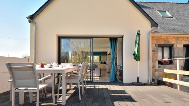 Una terraza de madera con una mesa y sillas, una puerta corrediza de vidrio que da a una cocina y un edificio de piedra con un ventanal.