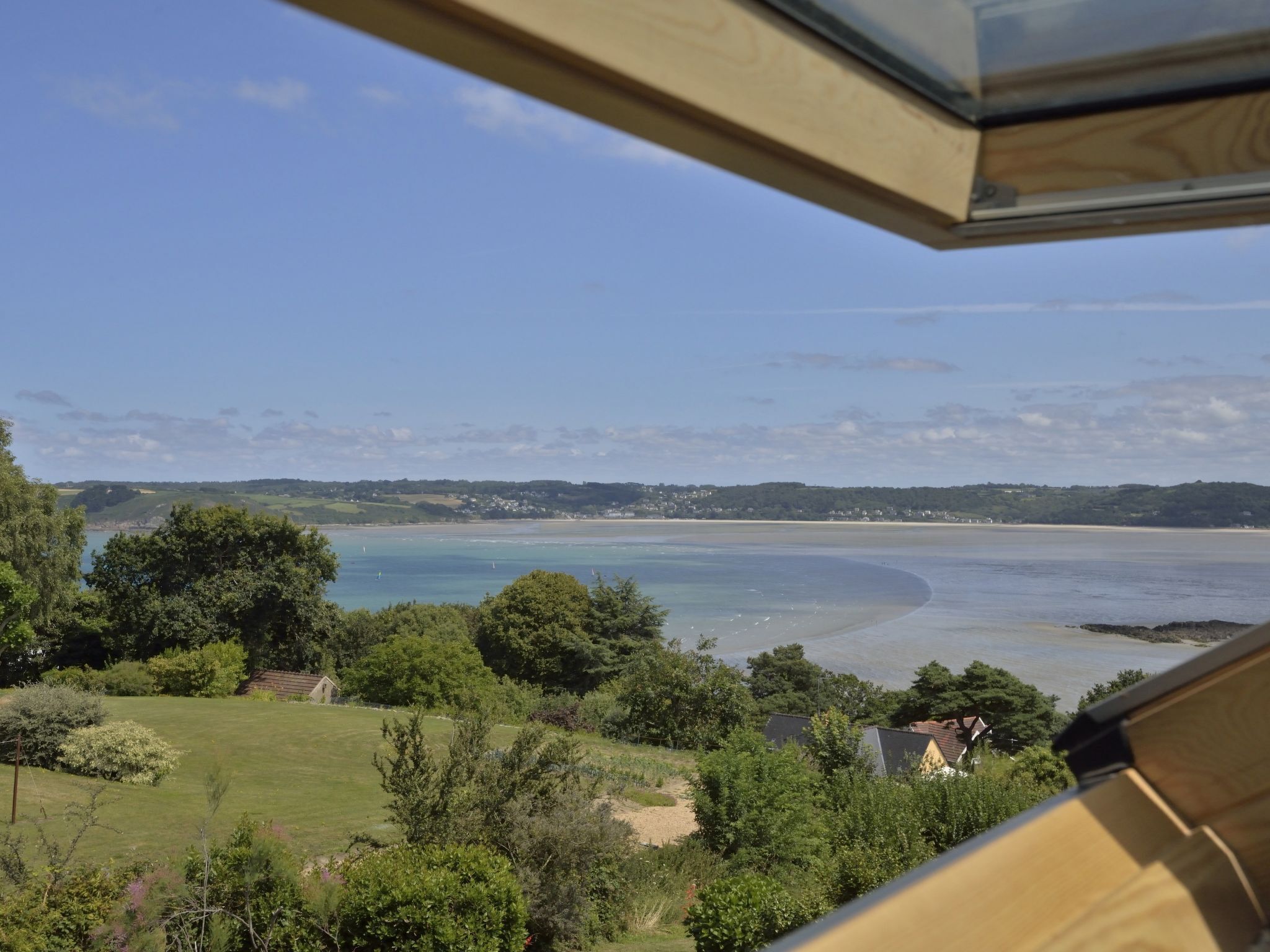 A coastal view from a room with a large window overlooking a beach, grassy areas, and a distant town.
