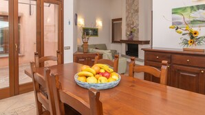 A dining area with a wooden table, chairs, and a fruit bowl. There is a stone fireplace, a television, and a sofa in the background. - Rei Sole (REI316) by Interhome (Costa Rei)