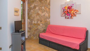A living room with a stone wall, a red sofa, a television on a stand, and a wall-mounted light fixture. - Rei Sole Bilo (REI321) by Interhome (Costa Rei)
