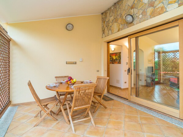 A dining area with a round table, wooden chairs, and a fruit bowl. There is a stone wall and a glass door leading to an outdoor area.