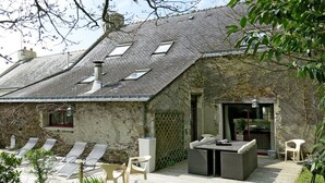 Una casa de piedra con techo inclinado, zona de estar exterior con sillas y una mesa, y una terraza de madera.