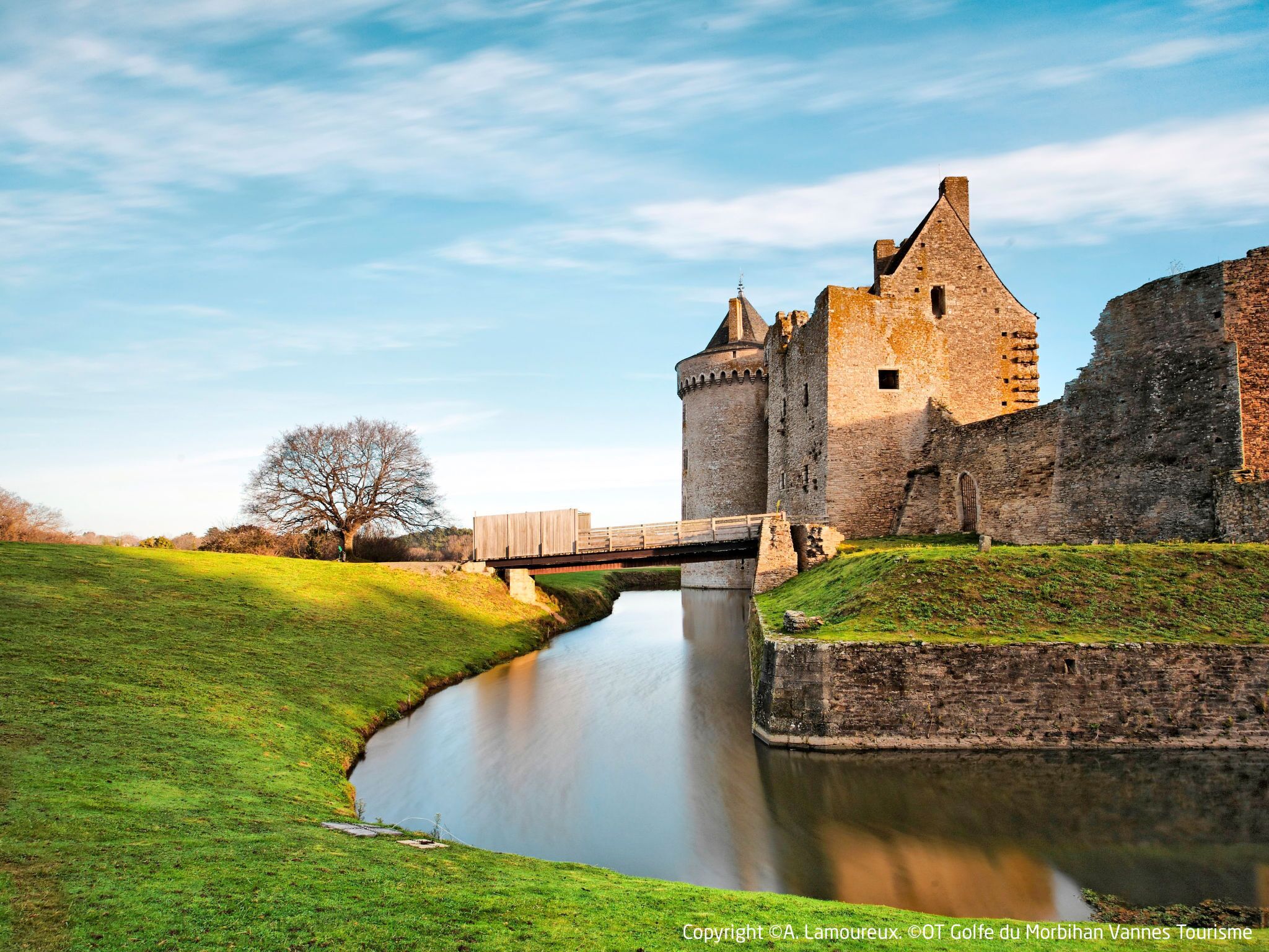A historic castle with a moat, a wooden bridge, and a grassy landscape.