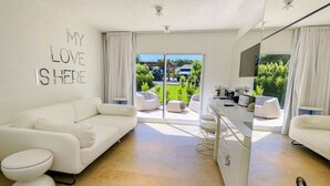 En moderne stue med en hvid sofa, et sofabord og et spiseområde med en køkkenø. Der er et stort vindue med gardiner, et loft med indbygningsbelysning og et skilt på væggen, der siger "MY LOVE IS HERE".