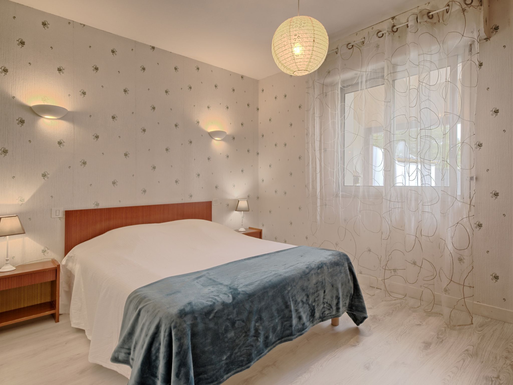 A bedroom with a bed, two bedside tables, a hanging light fixture, a window with sheer curtains, and patterned wallpaper.