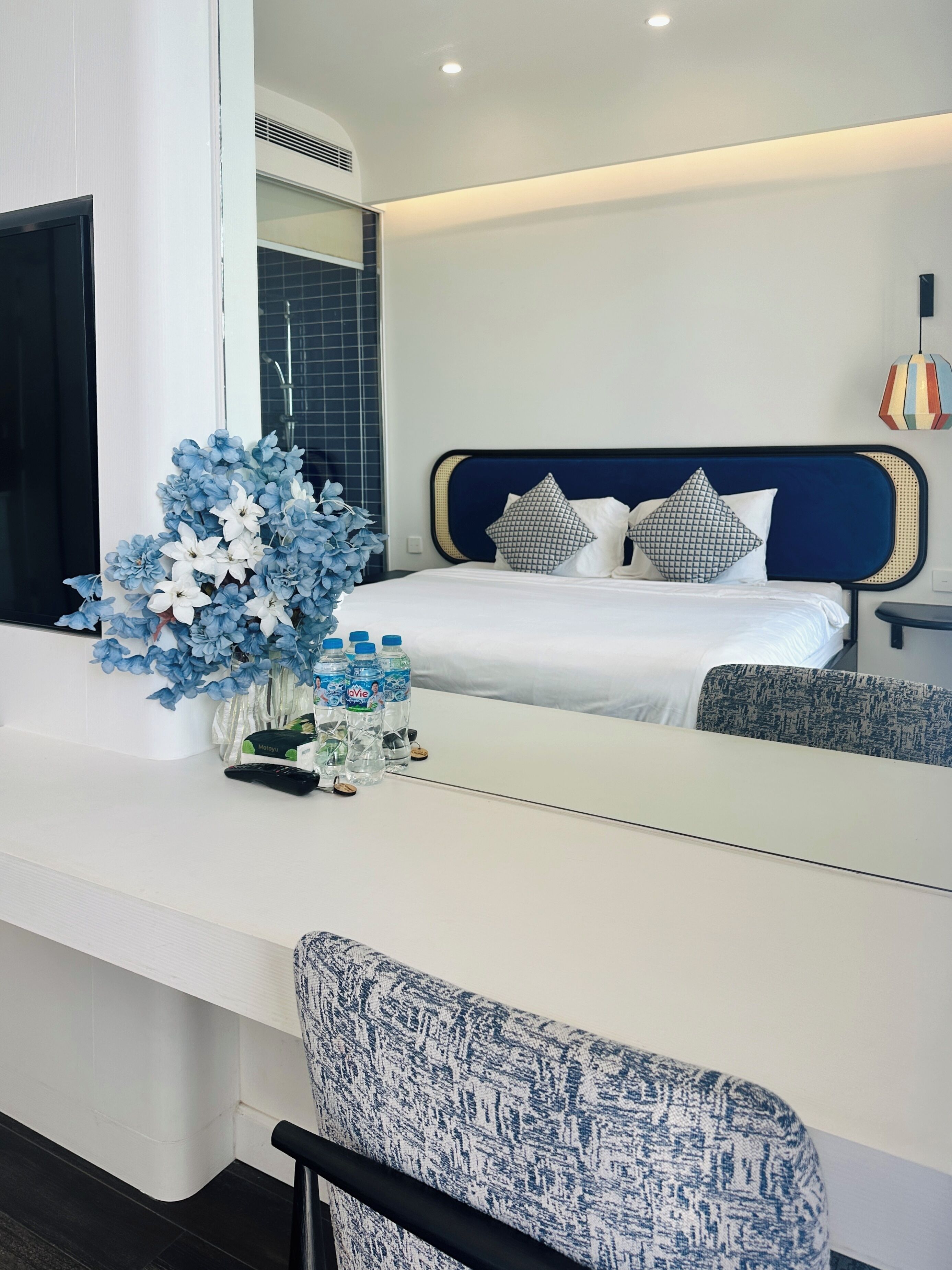 Une chambre d’hôtel moderne avec un grand lit, une table de chevet avec des bouteilles d’eau et un arrangement floral bleu décoratif.