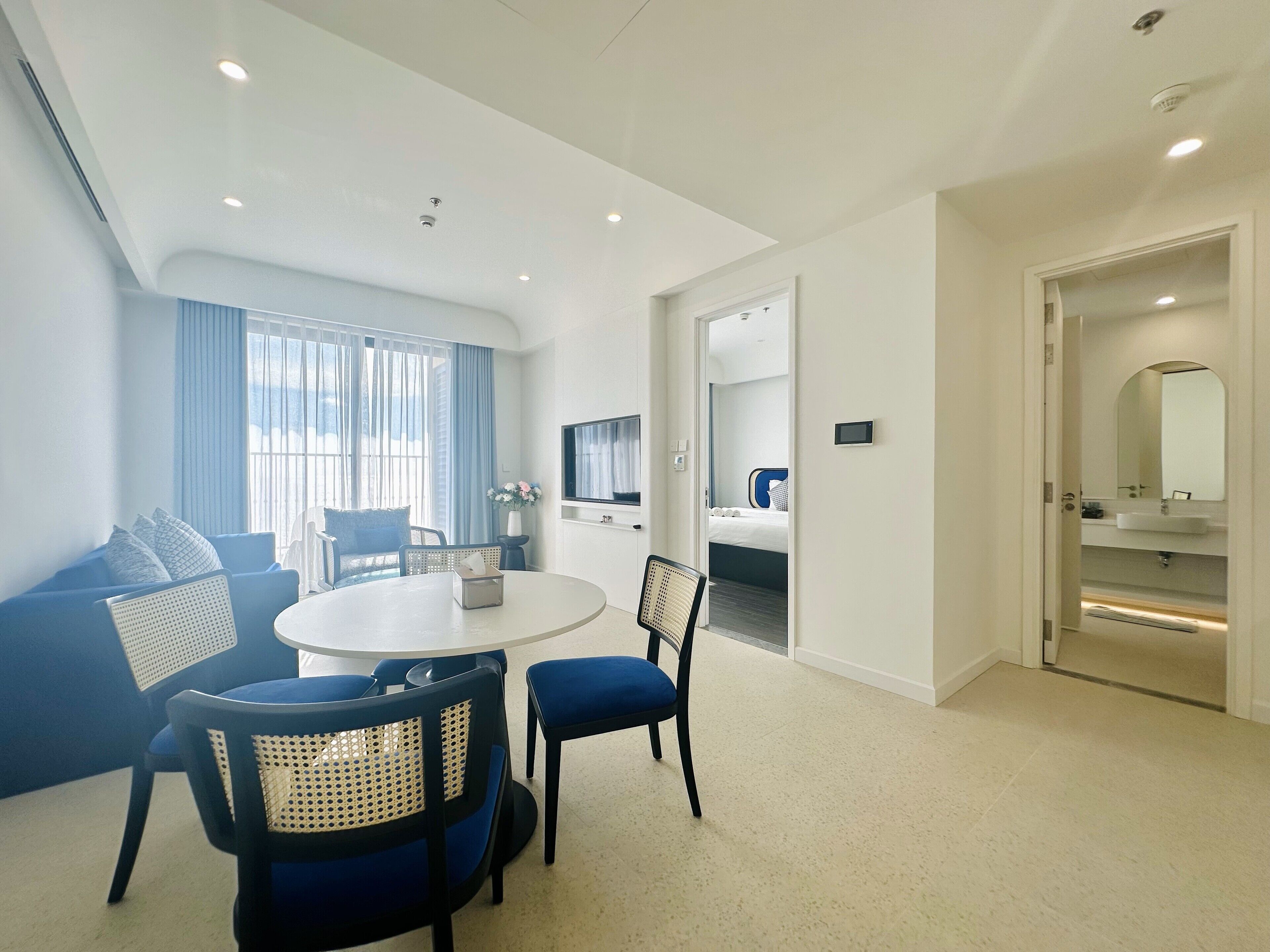 A modern hotel room with a round dining table, blue upholstered chairs, a sofa, a flat-screen TV, a bed with blue and white bedding, and a bathroom with a mirror and sink.