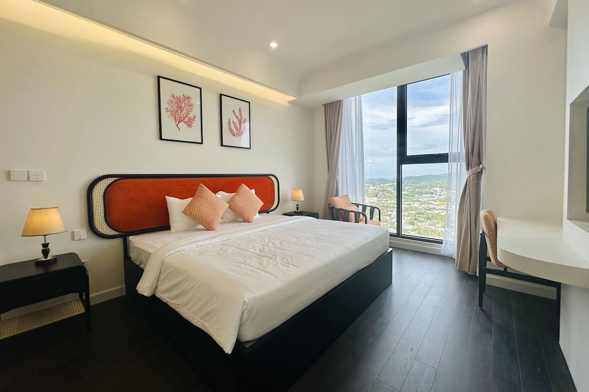 Une chambre d’hôtel moderne avec un grand lit, une table de chevet avec une lampe, une chaise et une grande fenêtre donnant sur la ville.