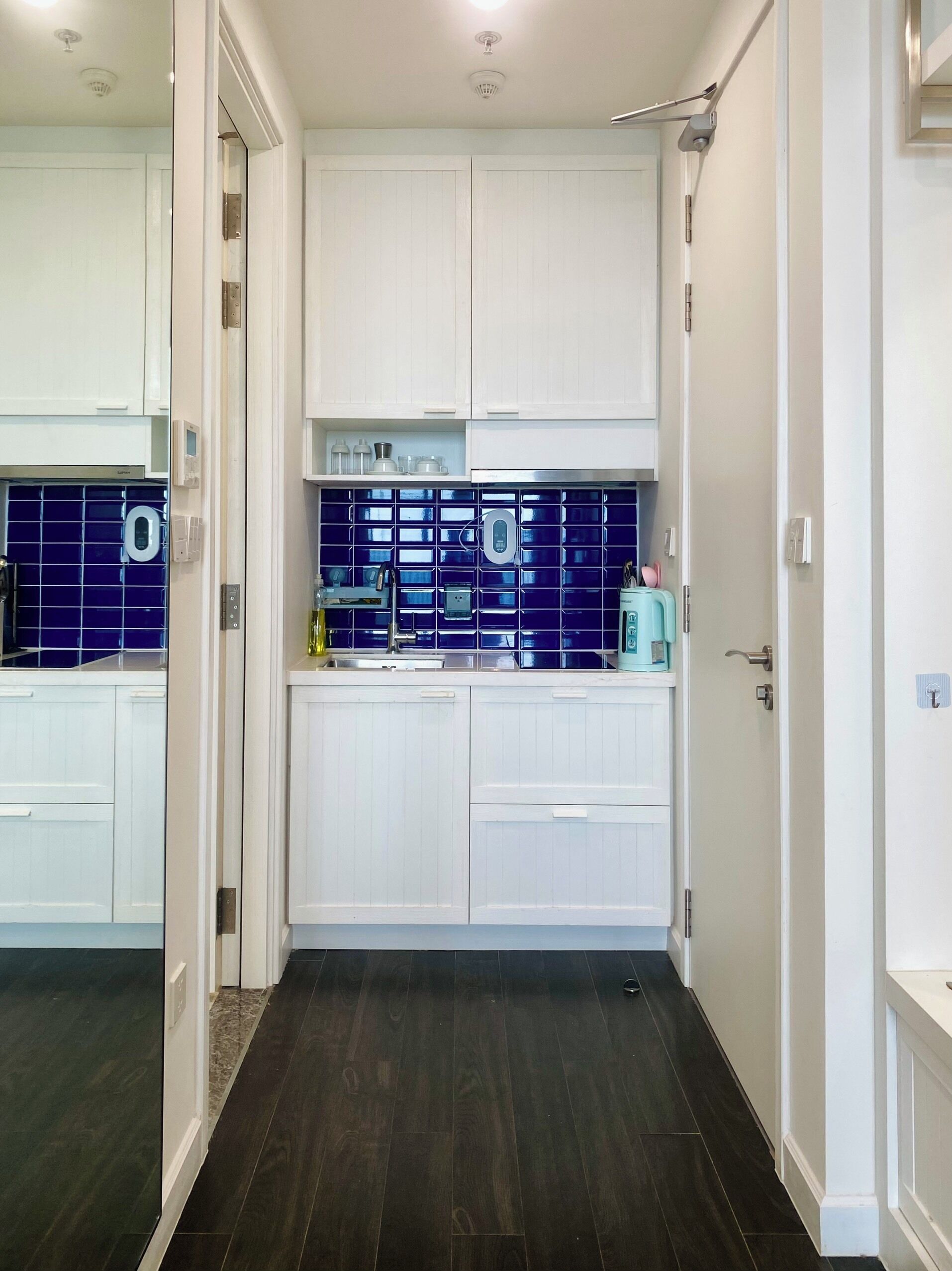 A narrow hallway with a dark wooden floor leading to a kitchen area with white cabinets and blue-tiled backsplash.