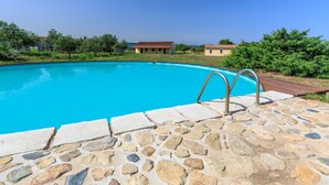 A swimming pool with a stone deck, surrounded by greenery and buildings in the background. - Loft la Pietraia by Interhome (Badesi)