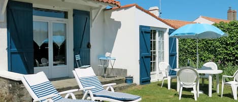 Un bâtiment blanc avec un toit en tuiles, des volets bleus et une antenne parabolique. On y trouve deux chaises longues avec des coussins à rayures et une table avec des chaises.