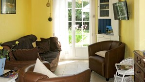 Een gezellige woonkamer met een bruine leren bank, een fauteuil en een klein tafeltje met tijdschriften. Een raam met gordijnen en zicht op groen is te zien.