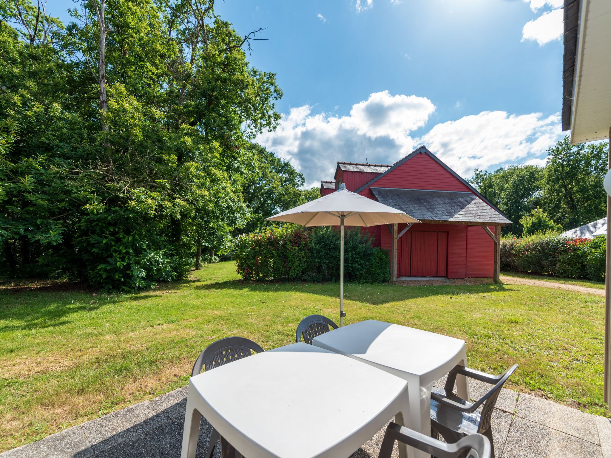 A patio area with a table and chairs, a red building in the background, and lush greenery.