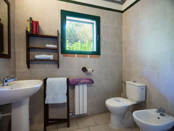 Un baño con lavamanos, inodoro, bidé y ventana con vistas a la vegetación.