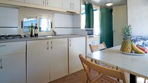 A compact kitchen with white cabinets, a microwave, and a window with curtains. A dining table with chairs is visible, and a fruit bowl with a pineapple, banana, and apple is on the table. - Campeggio del Forte by Interhome (Marina di Bibbona)