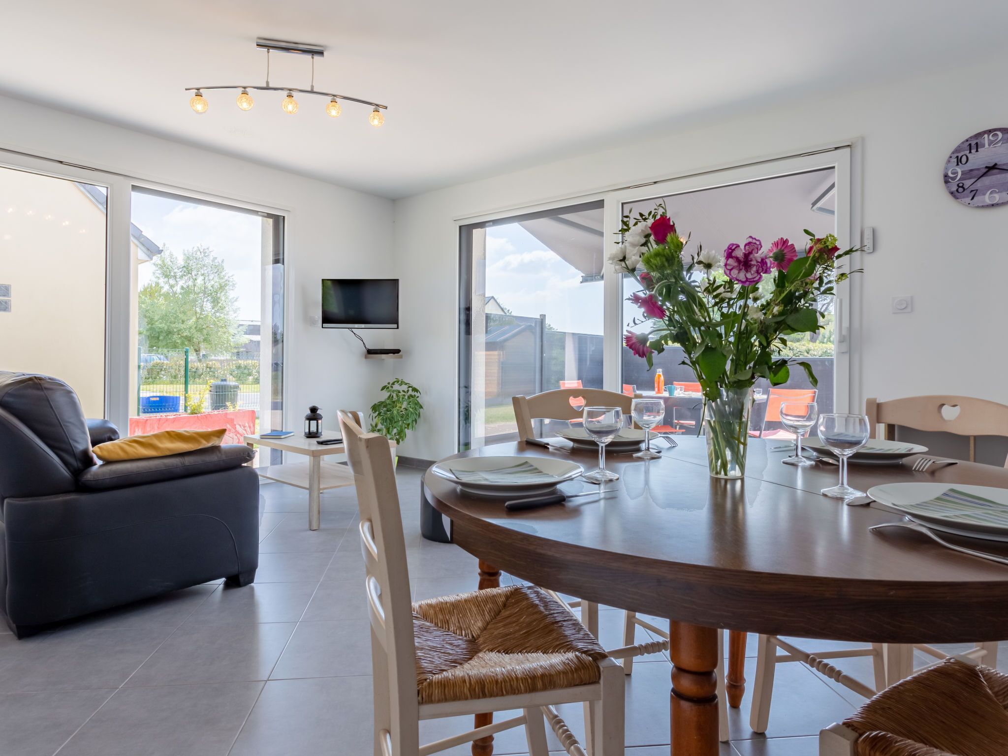A modern dining area with a round table, chairs, and a vase of flowers. There is a sofa and a TV mounted on the wall.