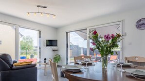 Ein moderner Essbereich mit einem runden Tisch, Stühlen und einer Blumenvase. Ein Sofa und ein an der Wand befestigter Fernseher sind ebenfalls vorhanden.