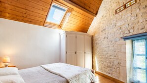 Una habitació amb sostre de fusta, paret de pedra, un llit amb llençols de retalls, un armari i una finestra amb cortina.