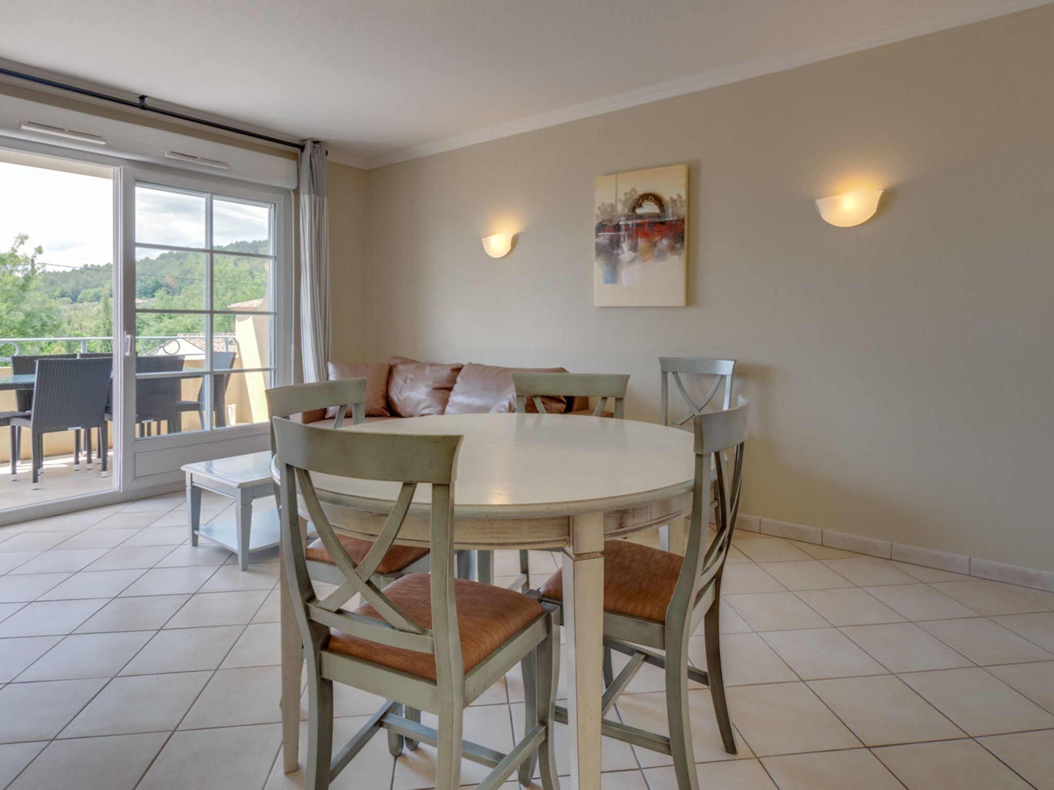 Uma área de refeições com uma mesa redonda, quatro cadeiras e um sofá. Há duas luminárias fixadas na parede e um quadro acima do sofá.