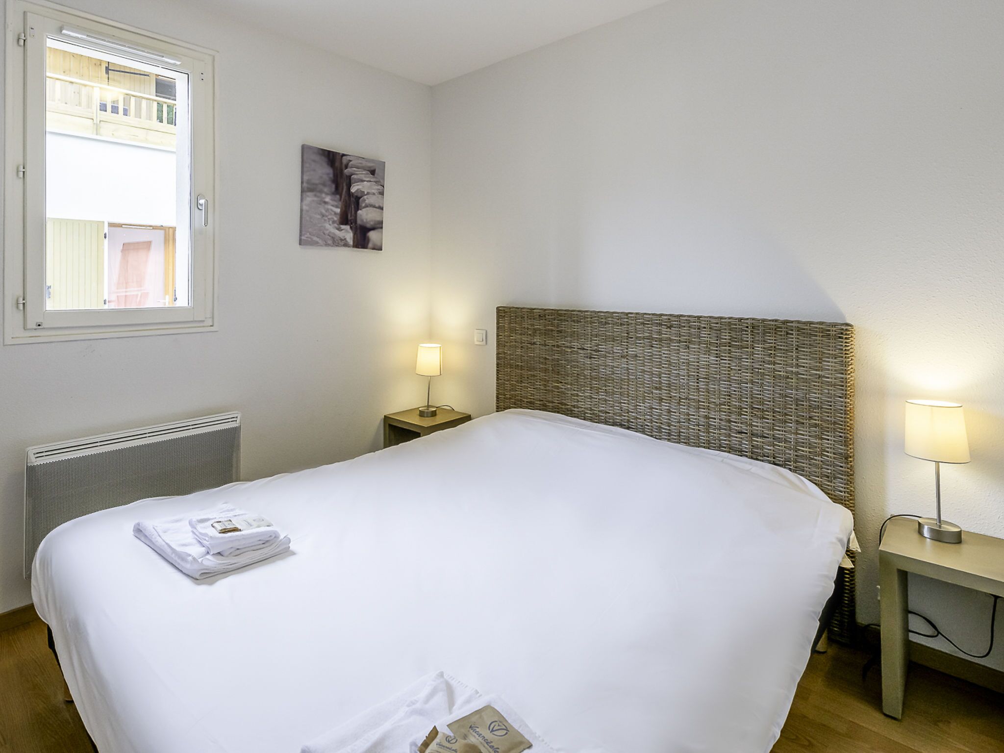 Een net opgemaakt bed in een hotelkamer, met een hoofdeinde, een nachtkastje met een lamp, een radiator en een raam met uitzicht op een gebouw.