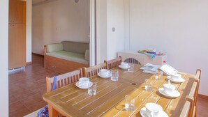 A dining area with a wooden table set for four, a sofa in the background, and a kitchenette with a stove and cabinets.