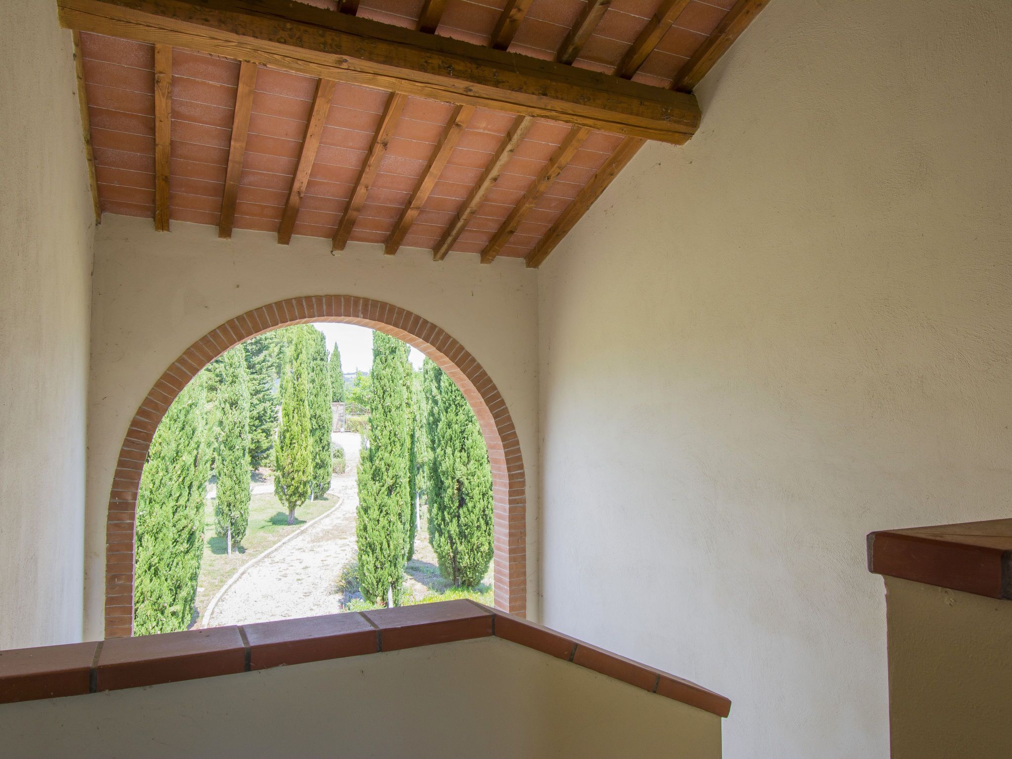 A view through an arched window with a brick arch and wooden beams above, leading to a pathway surrounded by tall trees.
