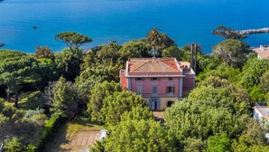 Uma villa costeira com telhado de telhas vermelhas, rodeada por vegetação exuberante e com vista para um mar tranquilo.