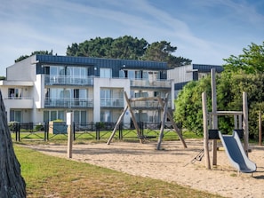 A playground with swings and a slide, surrounded by a fence, with a building in the background. - Odalys Domaine des Roches Jaunes by Interhome (Plougasnou)