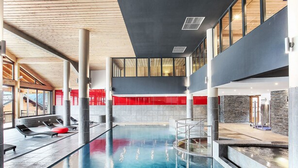 Une piscine intérieure moderne, avec de l’eau cristalline, des poutres en bois au plafond et des touches de couleur rouge.