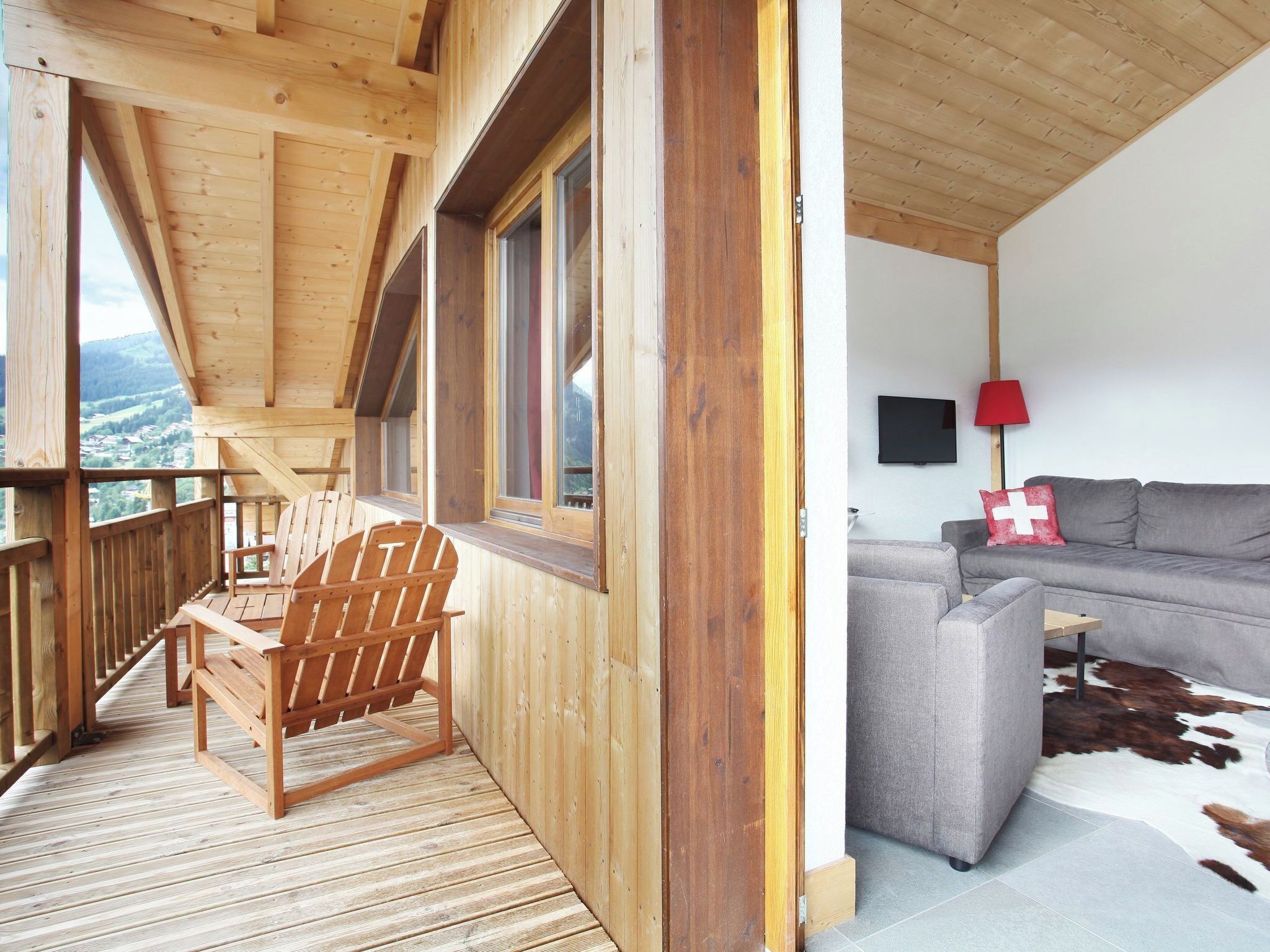 Un balcon en bois avec une chaise en bois, un canapé gris et un tapis en peau de vache, dans une pièce équipée d’un téléviseur et d’une lampe rouge.