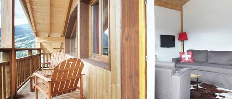 Un balcon en bois avec une chaise en bois, un canapé gris et un tapis en peau de vache, dans une pièce équipée d’un téléviseur et d’une lampe rouge.